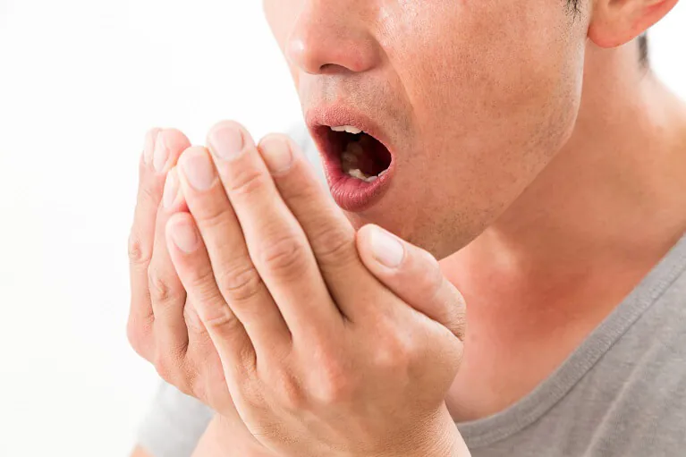 白い背景をバックに、自分の口臭を確認しようとする男性のクローズアップ画像。男性は口を大きく開け、両手のひらを顔の前にカップ状に添えて、吐き出した息の臭いをセルフチェックしている様子。