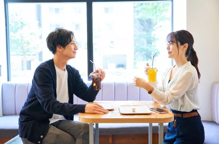 明るいカフェの店内で、木製のテーブルを挟んで向かい合い、飲み物を手に会話を楽しむ男女の様子。左側に座る男性は黒のカーディガンに白のTシャツを着用し、アイスコーヒーを手に穏やかな表情を浮かべています。右側に座る女性は白のブラウスとネイビーのパンツを身につけ、ポニーテールの髪型で、オレンジ系のドリンクを持ちながら男性を見つめています。