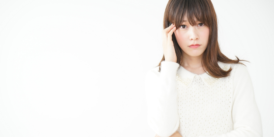 白を背景に、パールの装飾がついた白いニットを着た茶髪の女性が、右手でこめかみを押さえながら憂いのある表情を浮かべている様子。女性は視線を正面に向け、何か悩み事があるような、あるいは軽い頭痛や体調不良を感じているような、少し困った表情をしています。左側には大きな空白があり、清潔感のある白い空間の中に彼女の繊細な心理状態が際立つ、静かで情緒的な印象の画像です。
