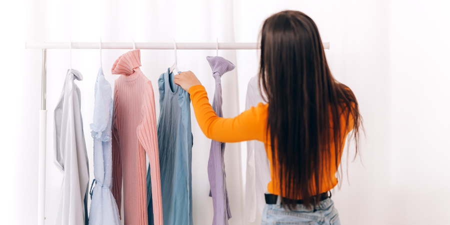 白を背景にした明るい室内で、オレンジ色の長袖トップスを着たロングヘアの女性が、ハンガーラックに掛かった服を選んでいる後ろ姿。ラックには、ピンクのタートルネック、水色のワンピース、薄紫のトップスなど、パステルカラーを中心とした多様なデザインの服が並んでいます。女性は右手を伸ばして青い服を手に取っており、外出の準備やショッピング、あるいは衣替えなど、日常のファッションを楽しむワクワク感や穏やかな時間が伝わってくる画像です。