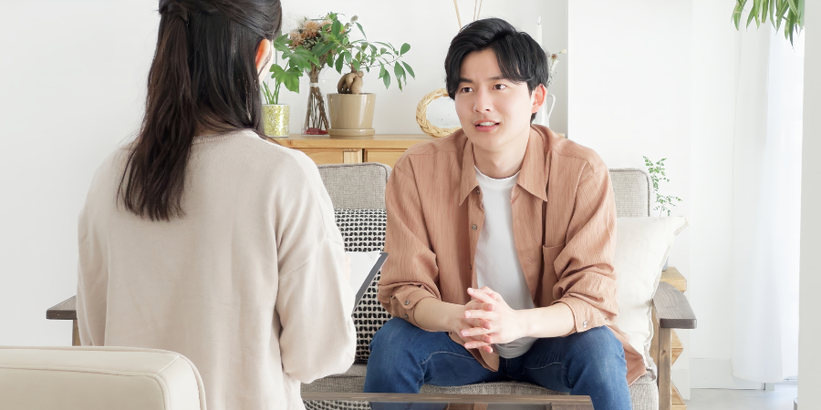 明るい室内で、グレーのソファに座る男性と、手前で背を向けて座る女性が向き合っている様子。茶色のシャツを着た男性は、少し困ったような、あるいは真剣に何かを相談しているような表情で、両手を組んで女性を見つめています。背景には観葉植物やナチュラルな木製の家具が配置され、落ち着いた雰囲気の中でのカウンセリングや、夫婦・パートナー間での真剣な話し合いを連想させる画像です。