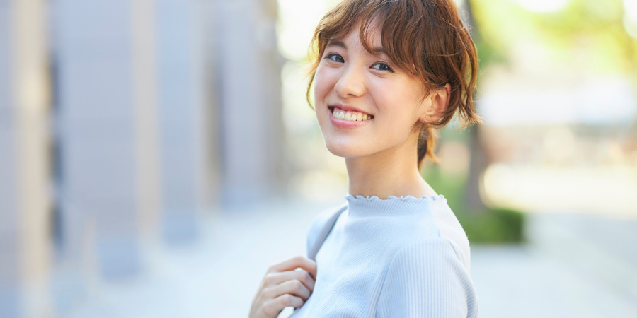 日差しが差し込む屋外で、明るい水色のトップスを着た若い女性が、肩にバッグをかけながら振り返り、カメラに向かって晴れやかな笑顔を見せている様子。ゆるくウェーブのかかった茶髪を後ろでまとめ、白く輝く歯を見せて笑う彼女の表情からは、自信とポジティブなエネルギーが感じられます。背景には街並みが柔らかくぼかして描かれており、日常の何気ない瞬間を切り取ったような、清潔感と親しみやすさが溢れる1枚です。