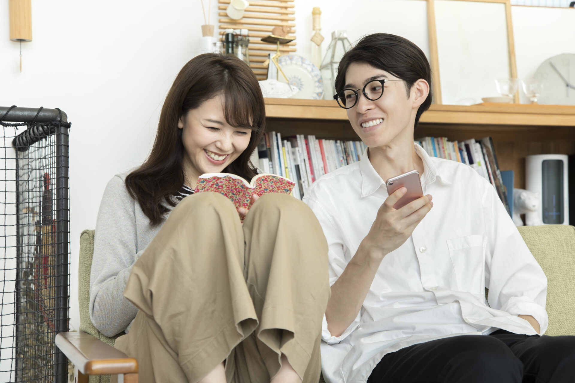 グリーンのソファに座り、リラックスした様子で過ごす男女の室内カット。左側の女性はグレーのニットにベージュのワイドパンツを合わせ、膝を抱えながら花柄のブックカバーがついた本を読み、楽しそうに笑っています。右側の男性は白のシャツに黒のパンツ、メガネを着用し、スマートフォンを手に持ちながら隣の女性を見て微笑んでいます。
