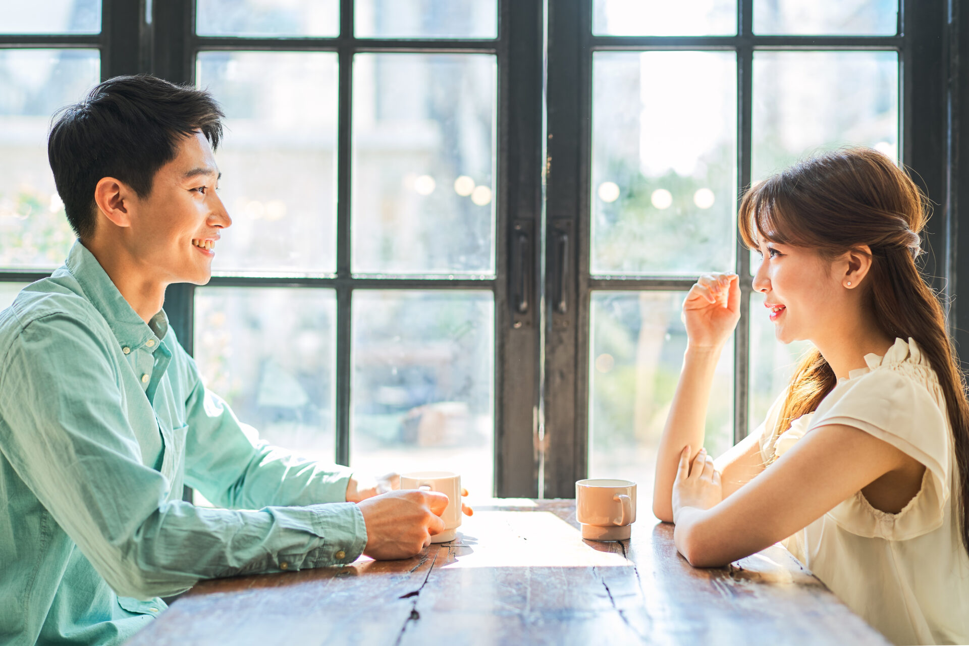 明るい光が差し込む大きな格子窓を背景に、木製のテーブルで向かい合って座る男女のクローズアップ。左側の男性はミントグリーンのシャツを着て、マグカップを手に持ちながら爽やかな笑顔で女性を見ています。右側の女性は白のノースリーブトップスを着用し、ハーフアップの髪型で、微笑みながら男性の話に耳を傾けています。