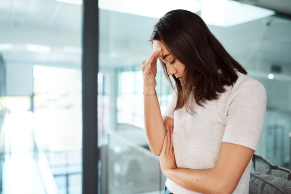 明るいオフィスのような場所で、白いTシャツを着た女性が沈痛な面持ちで立っている様子。女性は片手を額に当て、もう一方の手で自身の腕を抱えながら、視線を落として深く考え込んでいるような、あるいは疲れやストレスを感じているような表情を見せています。