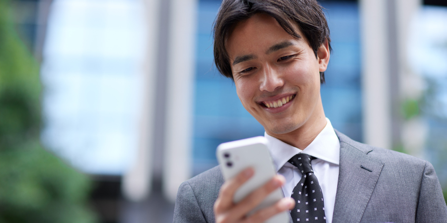 オフィス街の屋外で、スーツ姿の若い男性がスマートフォンを手に持ち、画面を見ながら爽やかに微笑んでいるアップの写真。