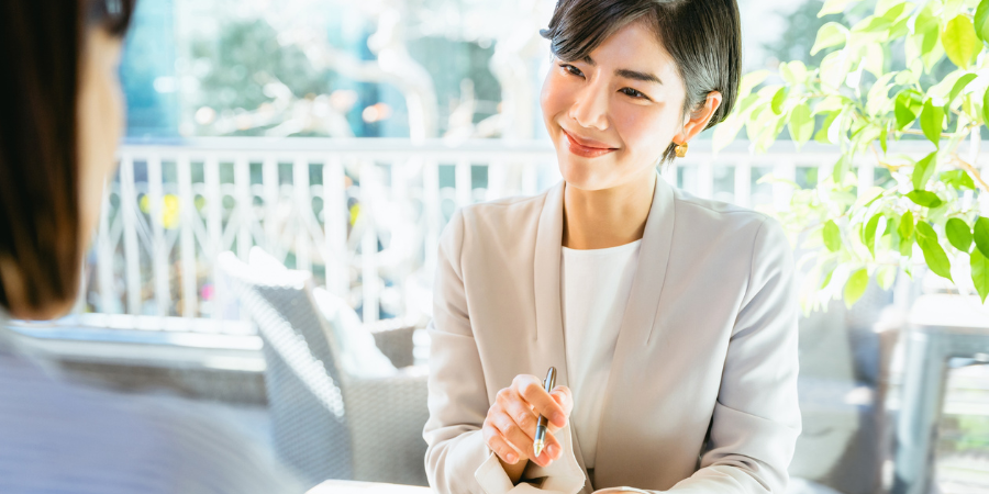 明るい屋外のテラス席で、手前に座る人物に向けて、ペンを持ちながら微笑みかけるベージュのジャケットを着たショートヘアの女性。