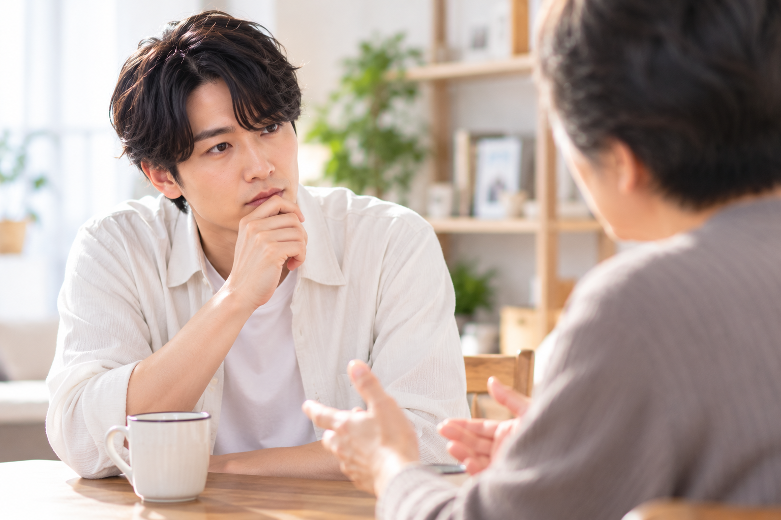自宅で親と向き合い、真剣に話を聞く若い男性。親の宗教と自分自身の結婚生活について考える様子を表現した写真風画像。