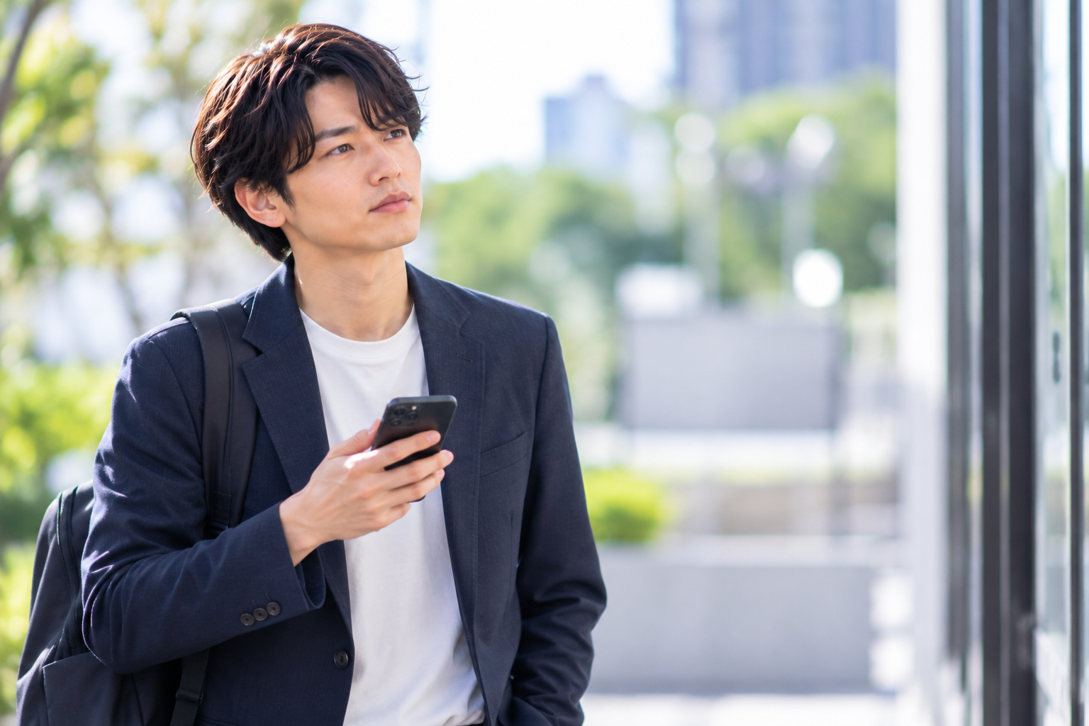 婚活中の男性が屋外でスマートフォンを手に、交際相手からの返事について考えている様子。親の宗教が結婚に与える影響や、交際終了への不安を表現した写真風画像。